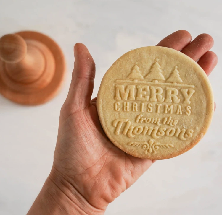 Dłoń trzyma świąteczne kruche ciasteczko z napisem „Merry Christmas”, wykonane drewnianym stemplem.