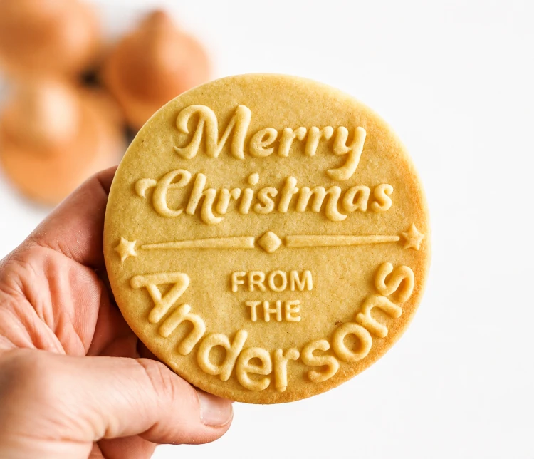 Personalizowane świąteczne ciasteczko z napisem „Merry Christmas from the Andersons” trzymane w dłoni. Wyraźny tłoczony wzór wykonany stemplem do ciastek z własnym nazwiskiem, idealny do bożonarodzeniowych wypieków i jadalnych prezentów.